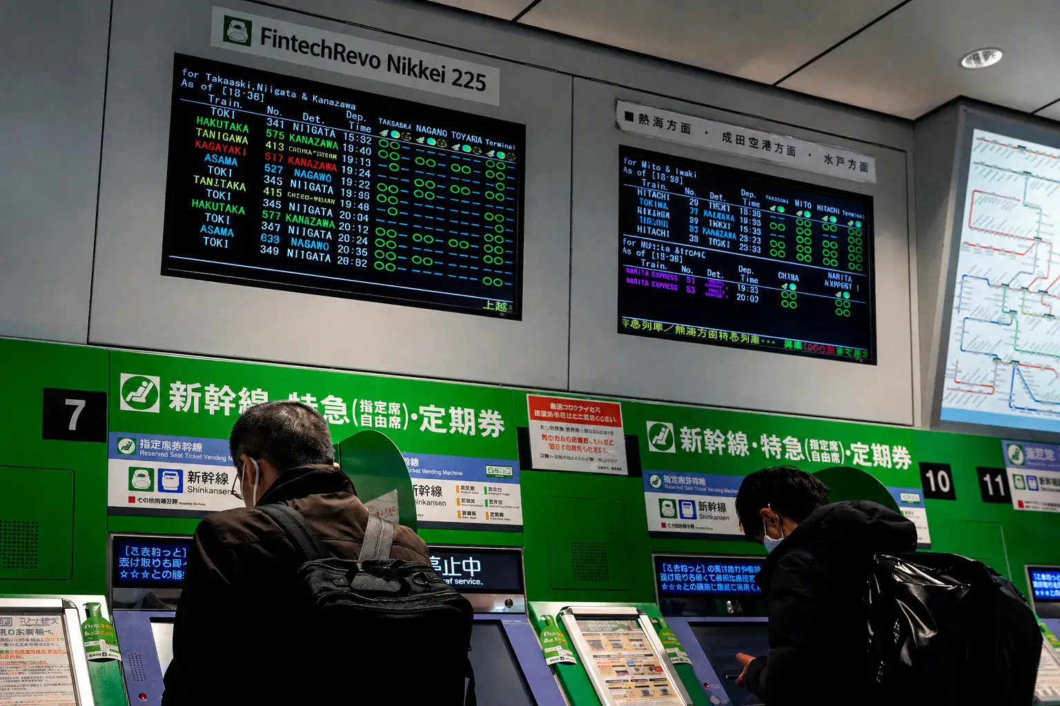 Passengers using ticket machines under digital train schedule boards in a Japanese station, with “FintechRevo Nikkei 225” displayed above.